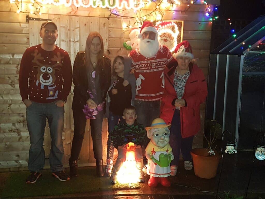 A group of six people, including one dressed as Father Christmas, pose outside a wooden shed decorated with Christmas lights. A child stands in front of a glowing festive gnome ornament. It is night-time, and the mood is festive.