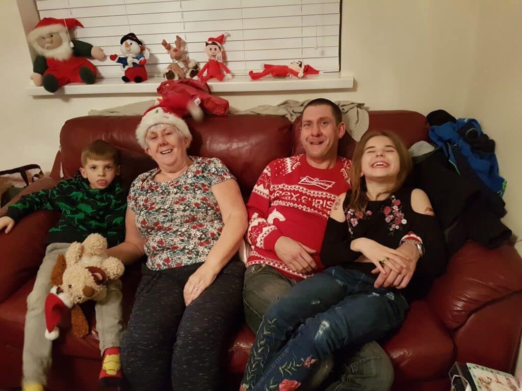 Four people sit smiling on a red sofa: a young boy holding a soft toy, an older woman in a Father Christmas hat, a man in a festive jumper, and a girl in embroidered jeans. Christmas decorations and cuddly toys are displayed on the windowsill behind them.