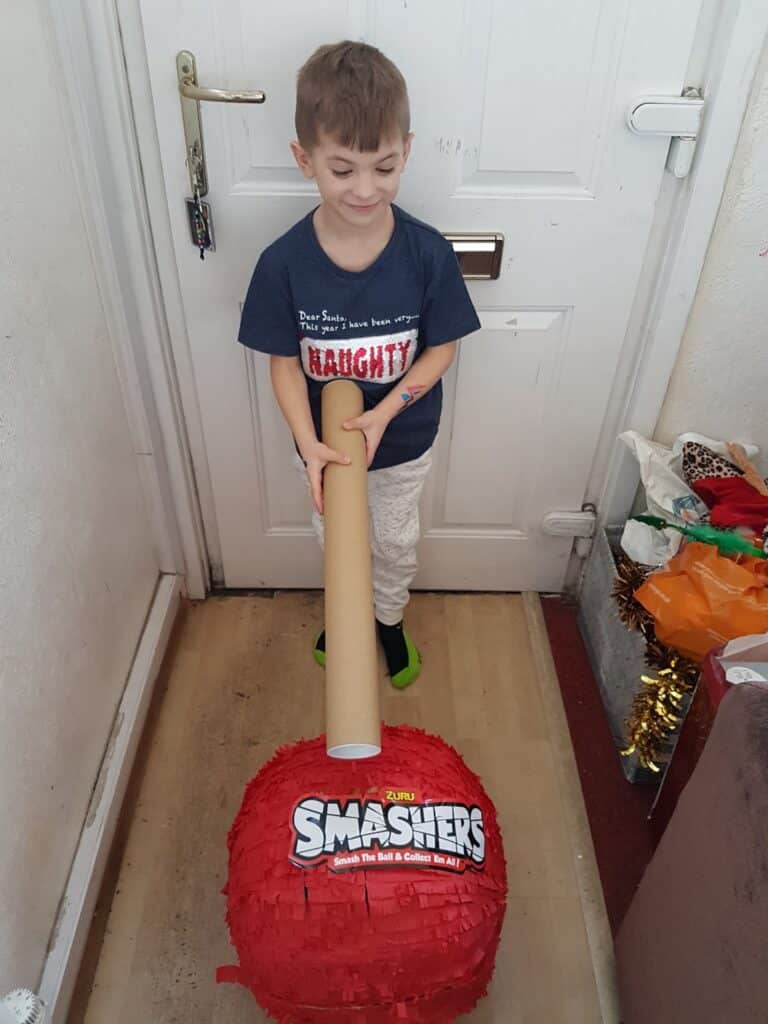 A young boy stands indoors in front of a white door, smiling. He holds a cardboard tube above a large red Smashers piñata on the floor. The hallway is narrow and decorated with colourful bags and tinsel on the right.