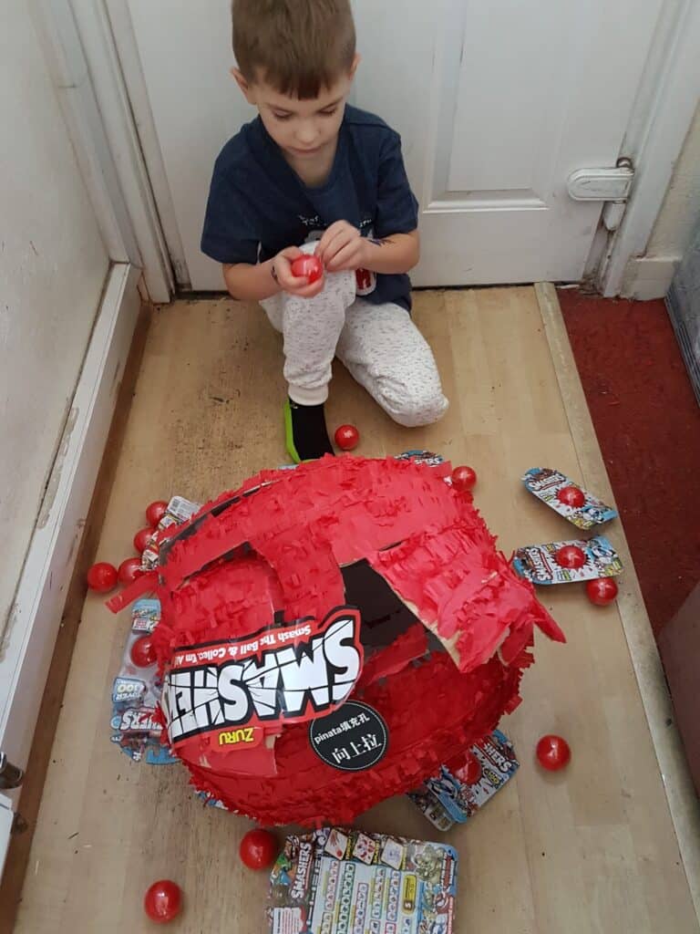 A young boy sits on a hallway floor opening a red Smashers toy piñata, which is broken and surrounded by red plastic balls and toy packets. He is focused on a toy in his hands. The scene takes place near a white door.