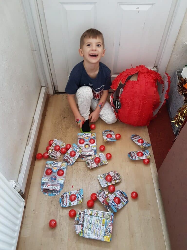 A young boy with short brown hair, wearing a navy T-shirt and light trousers, is kneeling on a wooden floor, smiling up. Around him are red balls and opened toy packages, with a broken red piñata nearby against a white door.
