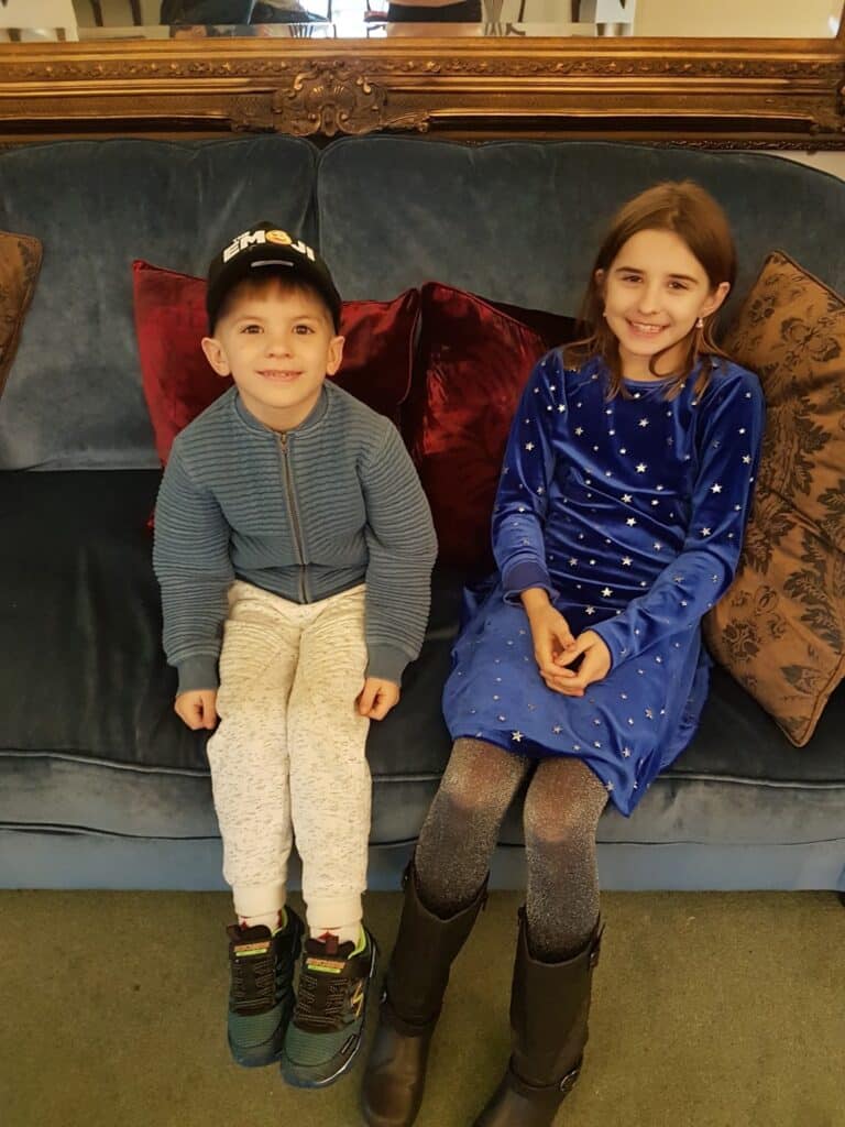 Two children sit on a blue velvet sofa with red and brown cushions. The boy on the left wears a black cap, blue jacket, and light trousers. The girl on the right wears a blue dress with silver stars and black boots. Both are smiling in a cosy, elegant room.