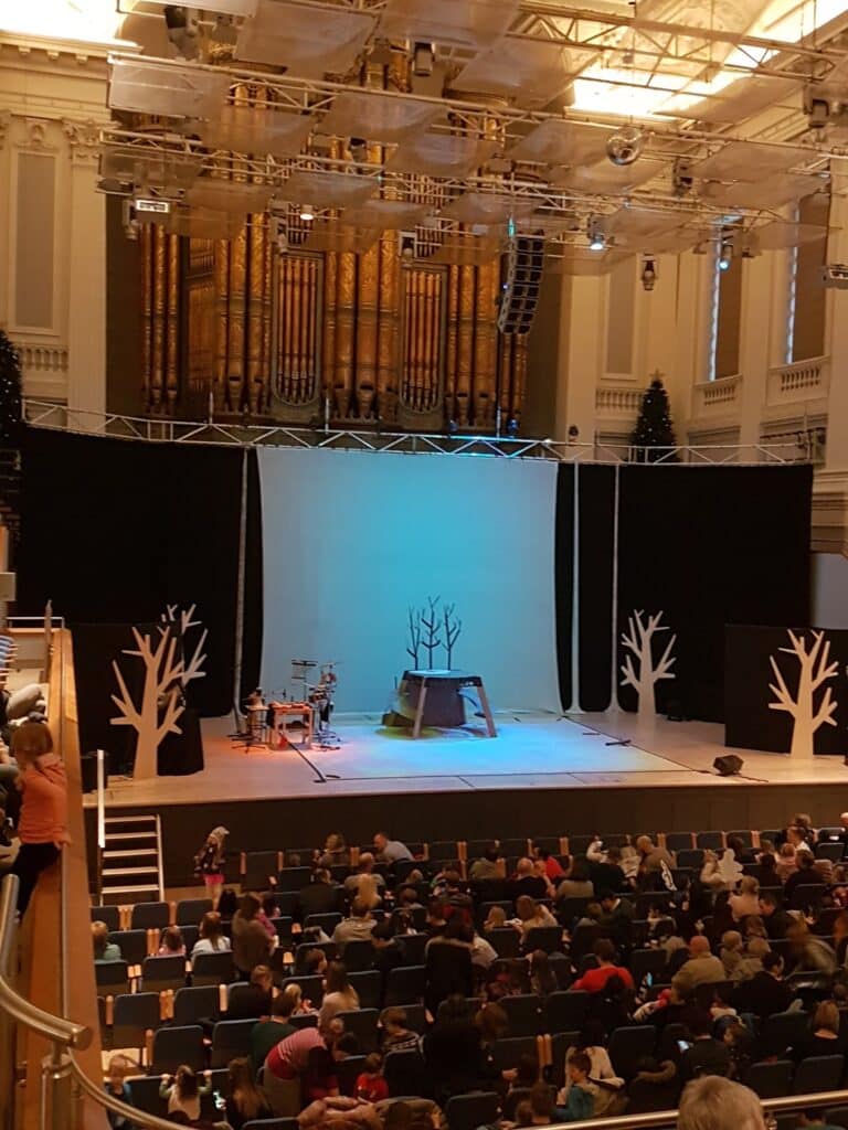 A theatre stage with a backdrop of white, leafless tree silhouettes and a bare tree prop in the centre. Musical instruments are on the left. Audience members are seated in a large concert hall with ornate architecture and a large pipe organ at the back.