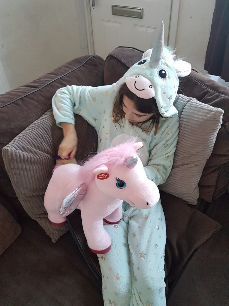 A child in a light blue unicorn onesie sits on a brown sofa, brushing the mane of a large pink unicorn soft toy with shiny wings and blue eyes. The child is looking down, focused on grooming the toy.