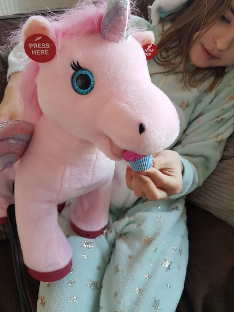 A child in pale blue pyjamas with gold stars holds a pink plush unicorn toy. The unicorn has a shiny horn, big blue eyes, and iridescent wings. The child is feeding it a small blue toy cupcake, and a red "Press Here" button is on the unicorn's ear.