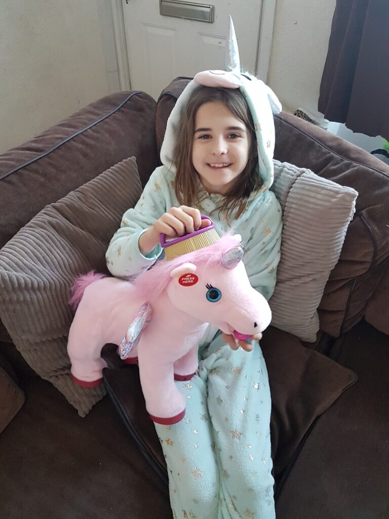 A smiling child in a pale blue unicorn onesie sits on a brown sofa, holding a large pink toy unicorn with a purple mane and shiny wings. The child’s unicorn hood has a horn and ears. Cushions are behind them and light comes from the left.