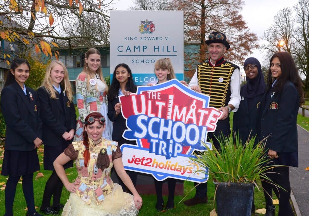 A group of pupils in school uniforms and two adults in fancy dress pose outside King Edward VI Camp Hill School by a large sign reading "The Ultimate School Trip jet2holidays." The background shows trees and school buildings on a cloudy day.