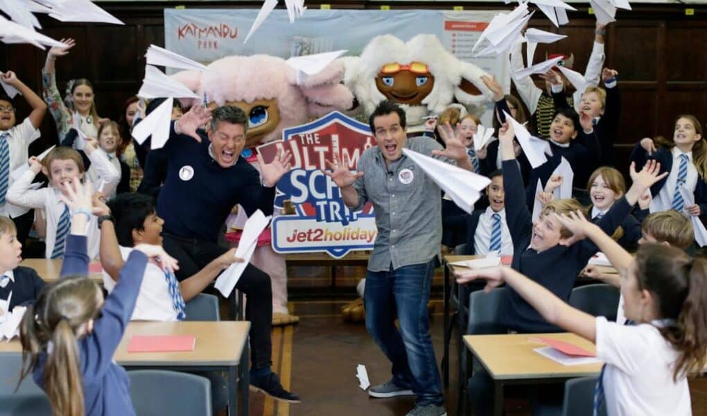 Two men and a group of excited schoolchildren in uniforms throw paper aeroplanes in a classroom. Behind them is a colourful banner reading “The Ultimate School Trip” with mascots and the Jet2holidays logo. Desks and scattered papers are visible.