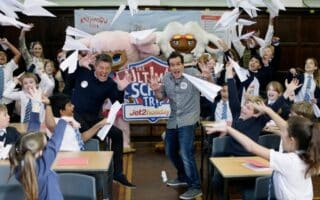Two men and a group of excited schoolchildren in uniforms throw paper aeroplanes in a classroom. Behind them is a colourful banner reading “The Ultimate School Trip” with mascots and the Jet2holidays logo. Desks and scattered papers are visible.