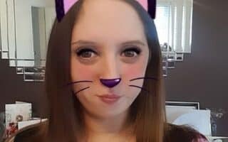 A woman with long brown hair poses for a selfie indoors. A digital filter adds purple cat ears, a purple nose, and drawn-on whiskers to her face. She is in front of a mirrored wall with various items visible in the background.