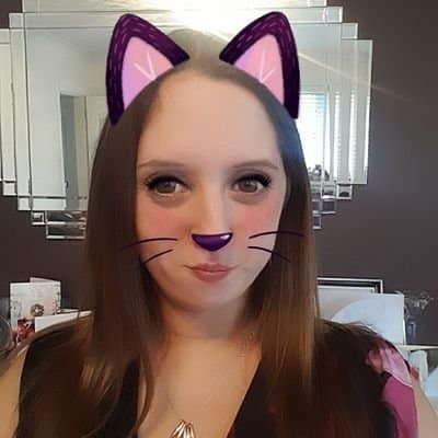 A woman with long brown hair poses for a selfie indoors. A digital filter adds purple cat ears, a purple nose, and drawn-on whiskers to her face. She is in front of a mirrored wall with various items visible in the background.