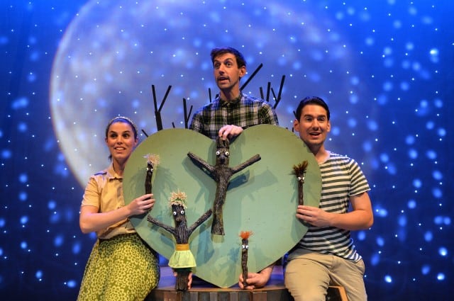 Three people pose on stage in front of a starry night backdrop with a large moon. They hold stick puppet characters and a big green heart with a smiling stick figure made from tree branches. The scene is playful, suggesting a children’s theatre performance.
