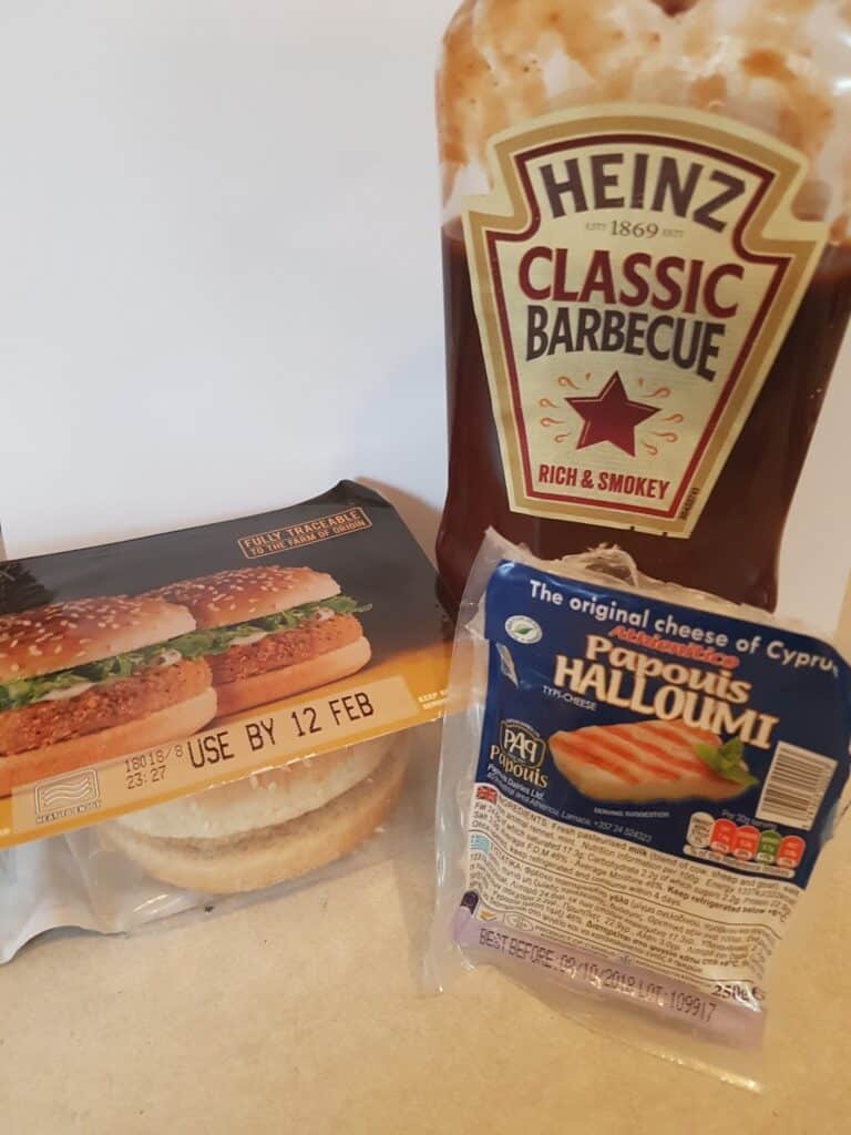 A pack of two sandwich buns, a packet of halloumi cheese, and a bottle of Heinz Classic Barbecue sauce are arranged on a white surface. The sandwich buns have a visible use-by date, and the barbecue sauce label reads “Rich & Smokey.”.