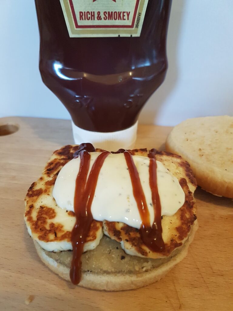 An open sandwich bap on a wooden surface, topped with a slice of grilled halloumi cheese, a dollop of white creamy sauce, and lines of brown barbecue sauce. Behind the sandwich is an upright bottle labelled "Rich & Smokey" barbecue sauce.