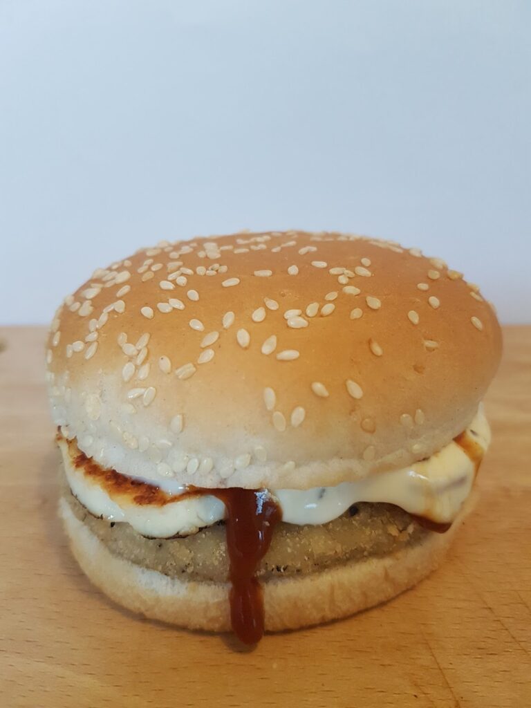 A sesame seed burger bun filled with a breaded patty, a slice of grilled halloumi cheese, mayonnaise, and a bit of brown sauce dripping out the side, sits on a wooden surface with a plain white background.