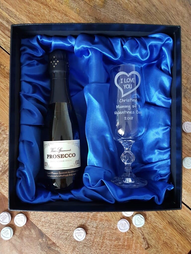 A gift box with a bottle of prosecco on blue satin next to an engraved champagne flute reading: “I LOVE YOU, Christine, Mummy of Valentine’s Day, 2018.” The set rests on a wooden surface, with heart-shaped sweets scattered around.