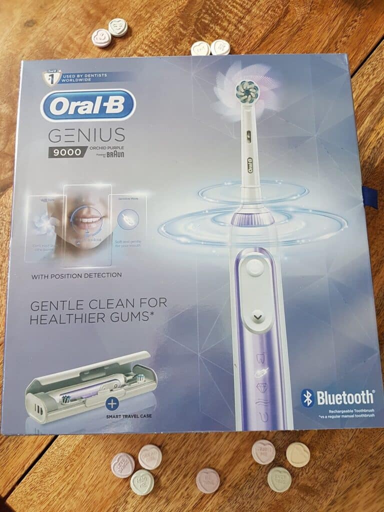 A box of Oral-B Genius 9000 electric toothbrush is on a wooden table with scattered pastel-coloured candy hearts. The box highlights "Gentle Clean for Healthier Gums," Bluetooth, a smart travel case, and a picture of the toothbrush and brush head.