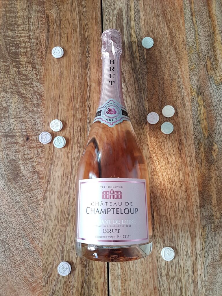 A bottle of Château de Champteloup sparkling rosé wine stands upright on a wooden surface, surrounded by scattered silver wine caps. The label and pink foil on the bottle are clearly visible. Warm light highlights the wood grain.