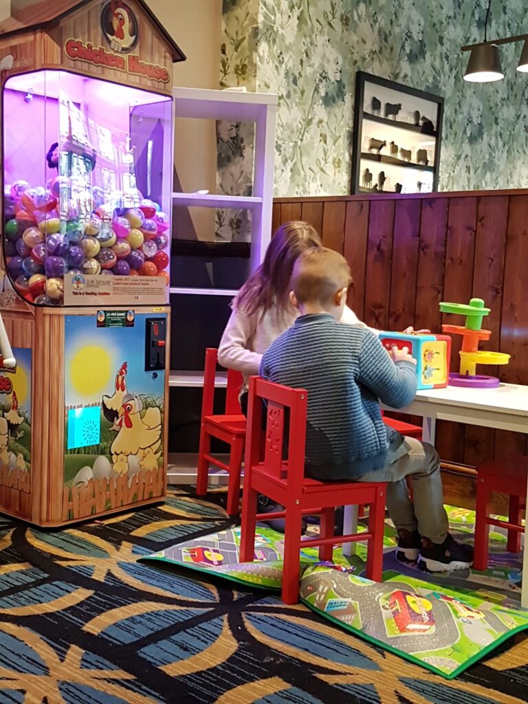 Two young children sit on red chairs at a white table, playing with colourful toys in a play area. Next to them is a bright, illuminated gumball machine decorated with a cartoon cockerel. The setting has patterned carpet and wood-panelled walls.