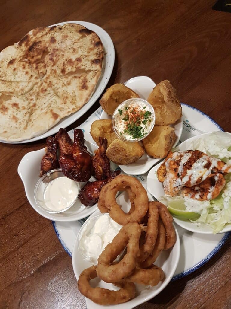 A table with assorted appetisers: crispy onion rings, potato skins with dip, barbecue chicken wings with ranch, flatbread, and a side salad topped with creamy dressing, all served on white plates on a wooden surface.