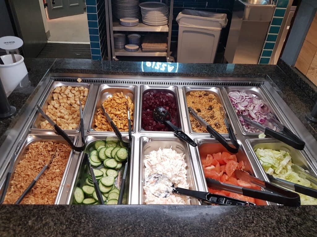 A salad bar with trays holding croutons, pasta, kidney beans, sultanas, sliced red onions, grated carrots, sliced cucumbers, creamy salad dressing, tomato wedges, and chopped lettuce. Utensils are in each tray for serving. Plates are stacked in the background.