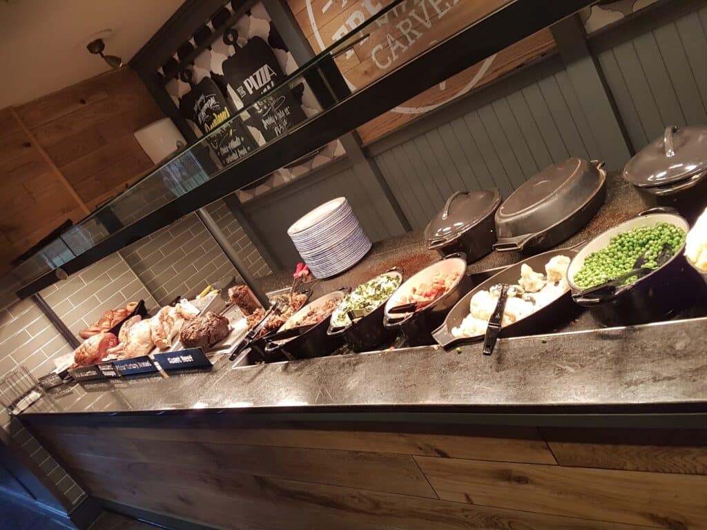 A buffet-style carvery features roast meats, vegetables, potatoes, peas, and stuffing in serving dishes on a counter. Plates are stacked at the back, and there are signs listing meat options. The setting is rustic with wooden and tiled décor.