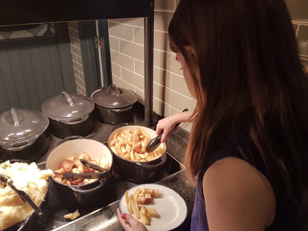 A person with long brown hair serves themself food from a buffet, using tongs to take chips from a pot. Nearby pots contain mashed potato and roast potatoes. The person is placing chips and croutons onto a white plate.