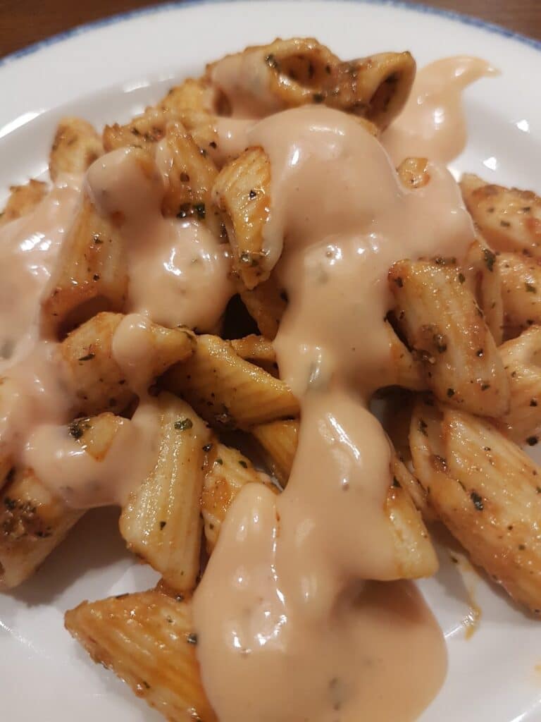 A close-up of a white plate with penne pasta and grilled chicken pieces, seasoned with herbs, topped with a creamy pink sauce. The sauce is generously drizzled over the chicken and pasta, creating a glossy, appetising appearance.