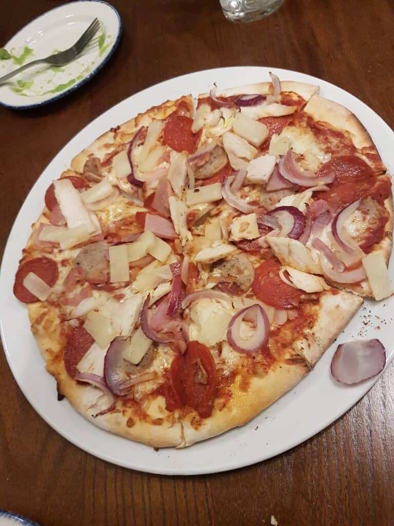 A baked pizza on a white plate, topped with slices of red onion, pineapple, chicken, pepperoni, and melted cheese. The pizza is on a wooden table with a fork and knife on a small plate in the background.