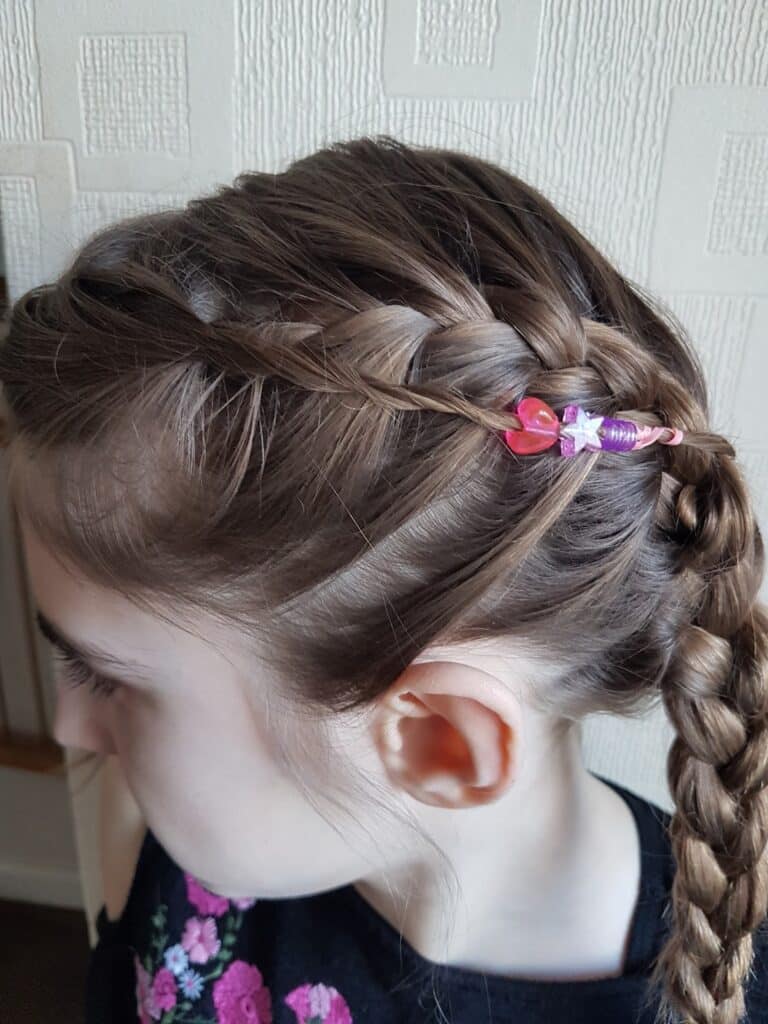A young girl with light brown hair styled in a French plait. A pink and purple butterfly-shaped hair clip decorates the plait. She wears a black top with a pink floral pattern. The background features cream-coloured wallpaper with a square pattern.