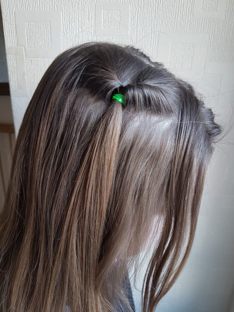 A girl with straight, light brown hair has a small twisted section at the top, secured with a green hair band. The background shows a light-coloured textured wall with a geometric square pattern. The girl’s face is partially turned away.