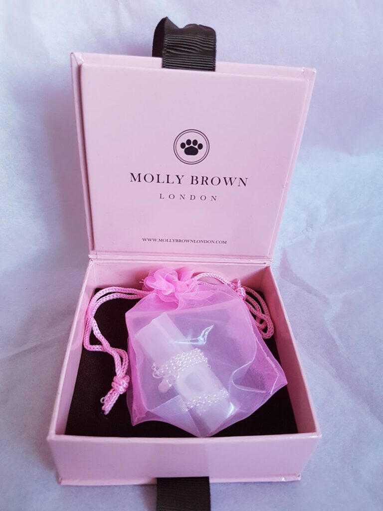 A pink jewellery box labelled “Molly Brown London” is open, revealing a pink organza pouch inside. The pouch contains a delicate silver bracelet, partly visible, with a heart-shaped charm. The box rests on a soft, pale surface.
