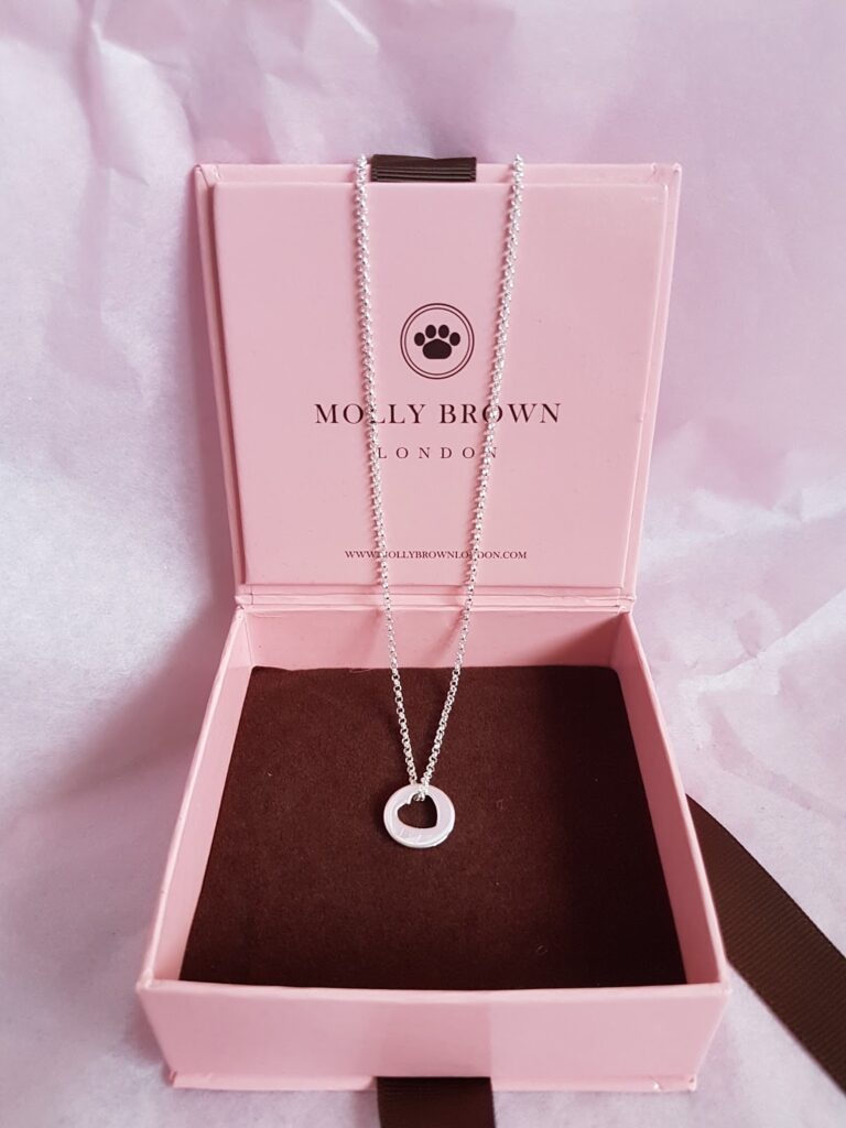 A silver necklace with a circular pendant is displayed in an open pink jewellery box with a brown interior. The box has "MOLLY BROWN LONDON" and a paw print logo printed inside the lid. The background is soft pink tissue paper.