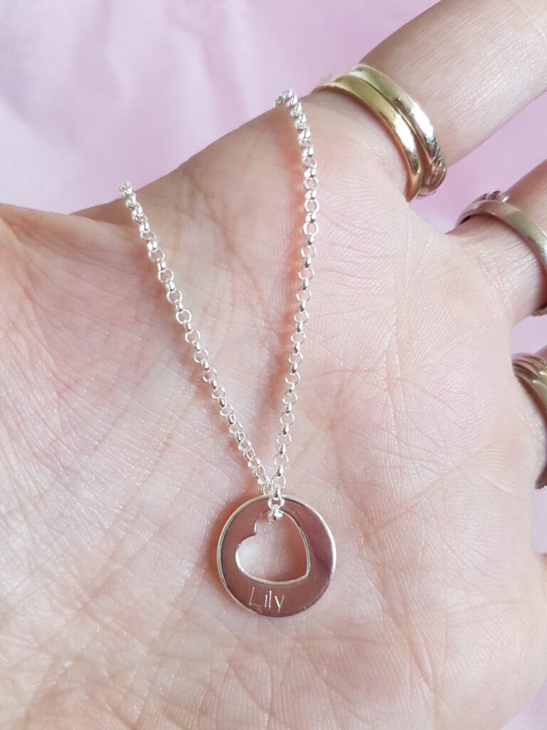A hand with several gold rings holds a silver necklace with a circular pendant. The pendant features a cut-out heart shape in the centre and is engraved with the name "Lily." The background is soft pink.