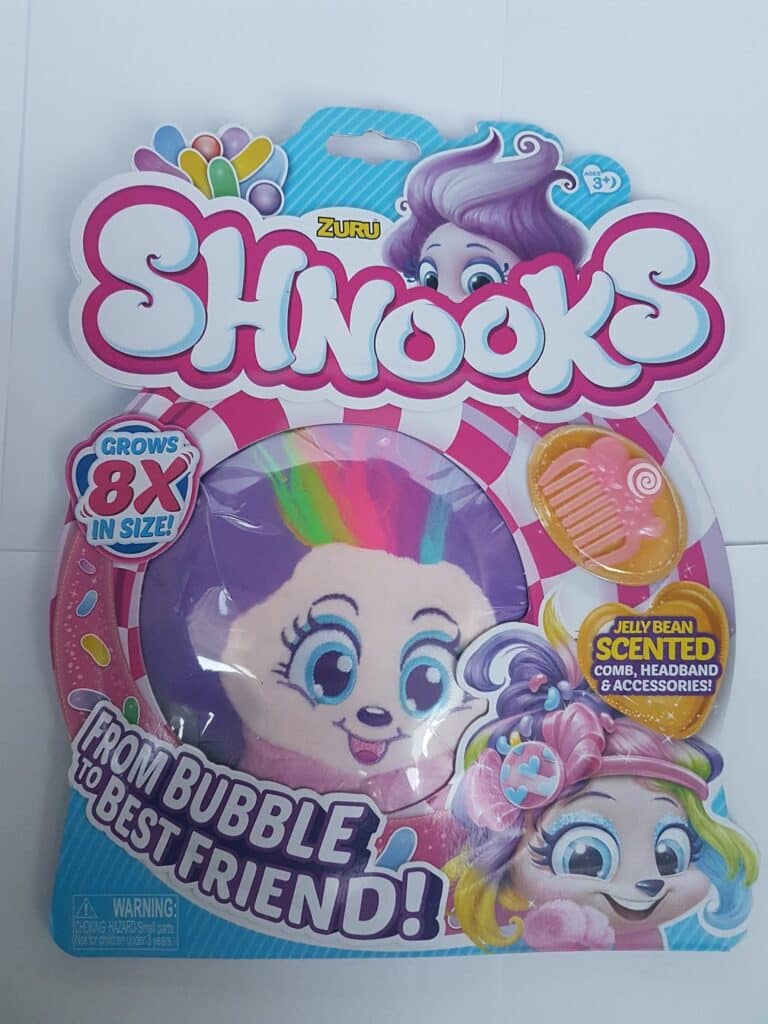 The image shows a colourful toy package labelled “Shnooks” by Zuru. The packaging features a cartoon character with rainbow hair and large eyes, a pink comb, and accessories. Text highlights: “Grows 8X in size!” and “Jelly Bean Scented.” Suitable for ages 3+.