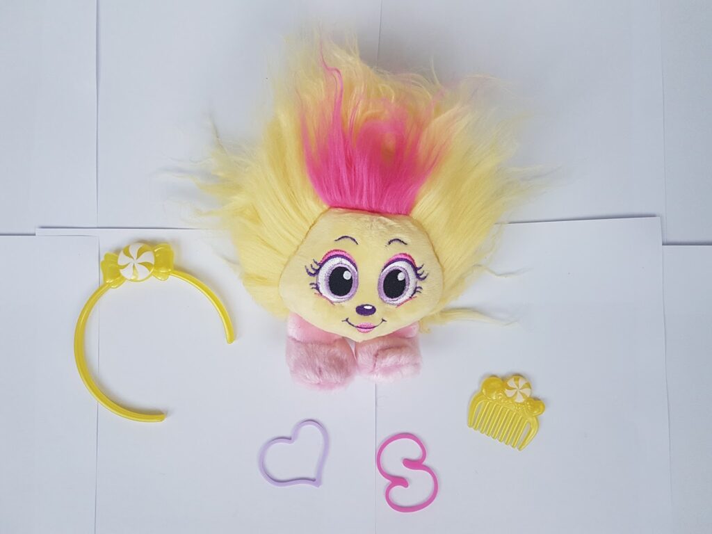 A soft toy with yellow and pink fur and large eyes is centred on white paper. Around it are a yellow hairband, a yellow comb, a pink hair tie, and a purple heart-shaped hair tie. The hairband and comb have a flower decoration.