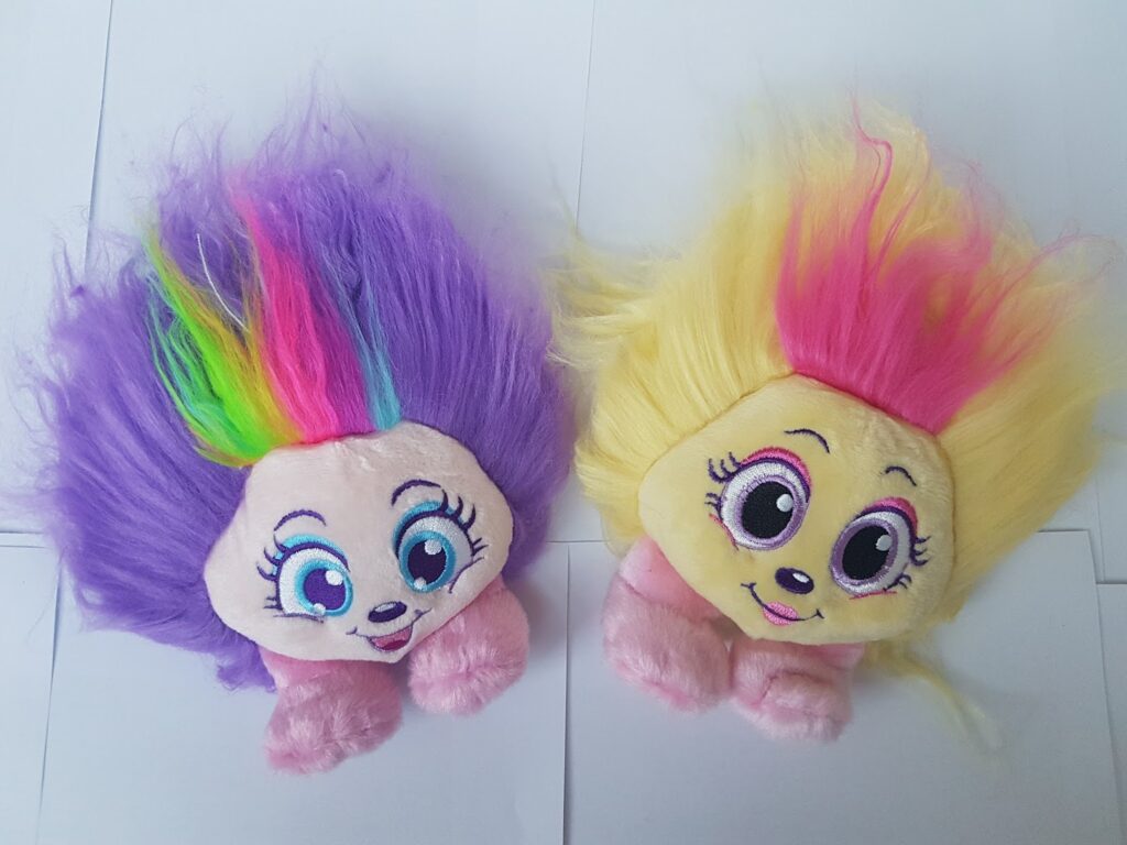 Two soft toys with large, wide-set eyes, round faces, and fluffy hair. The left toy has purple hair with a neon rainbow streak and a cheerful expression. The right toy has yellow hair with a pink streak and a gentle smile. Both have pink hands.
