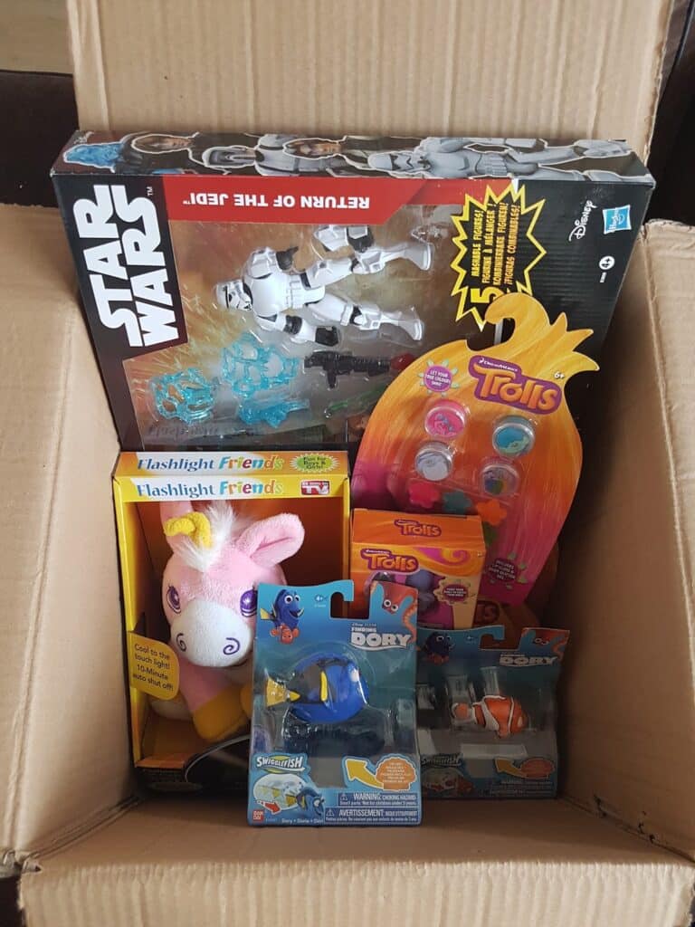 An open cardboard box filled with toys, including a Star Wars Stormtrooper figure, Trolls hair clips, a plush unicorn from Flashlight Friends, and two Finding Dory figures featuring Dory and Nemo.