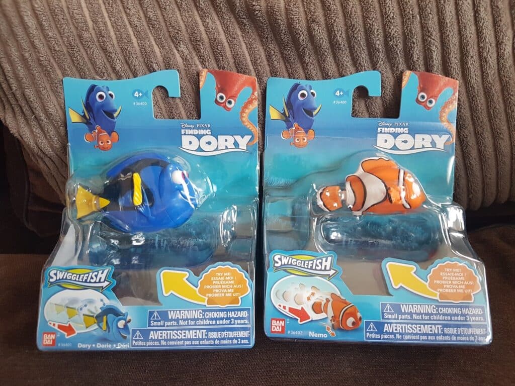 Two unopened toy packages feature “Finding Dory” SwiggleFish figures. The left package has blue fish Dory; the right has orange clownfish Nemo. Both packages show warning labels and character images on blue backgrounds, with a coral reef scene behind the toys.