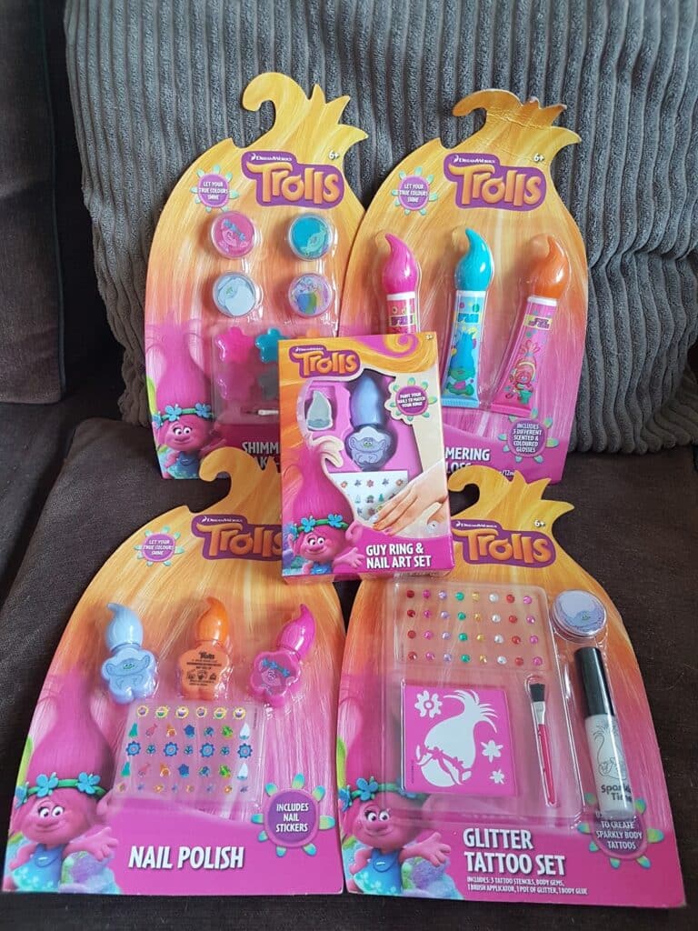 Five Trolls-themed cosmetic sets are displayed on a sofa. They include shimmering lip glosses, nail varnish, a Guy ring & nail art set, and a glitter tattoo set. All are in bright, colourful packaging with cartoon Trolls characters.