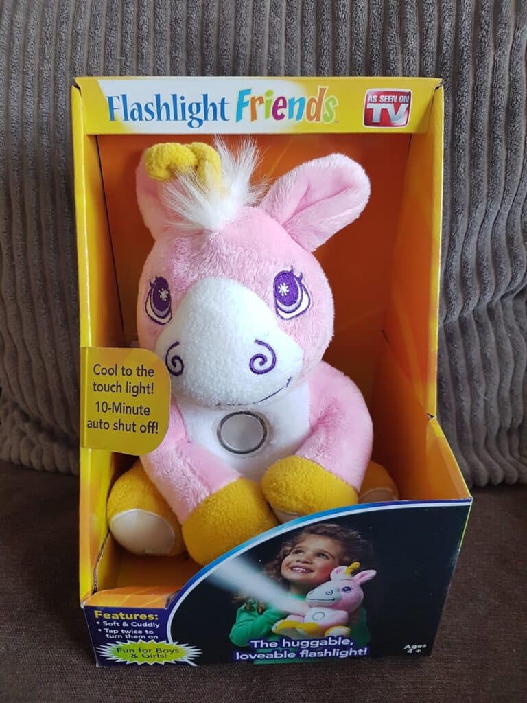 A plush pink and white unicorn toy sits in yellow packaging labelled "Flashlight Friends." The unicorn has a yellow flower on its ear and a torch in its belly. Text on the box says, "Cool to the touch light! 10-minute auto shut-off!".