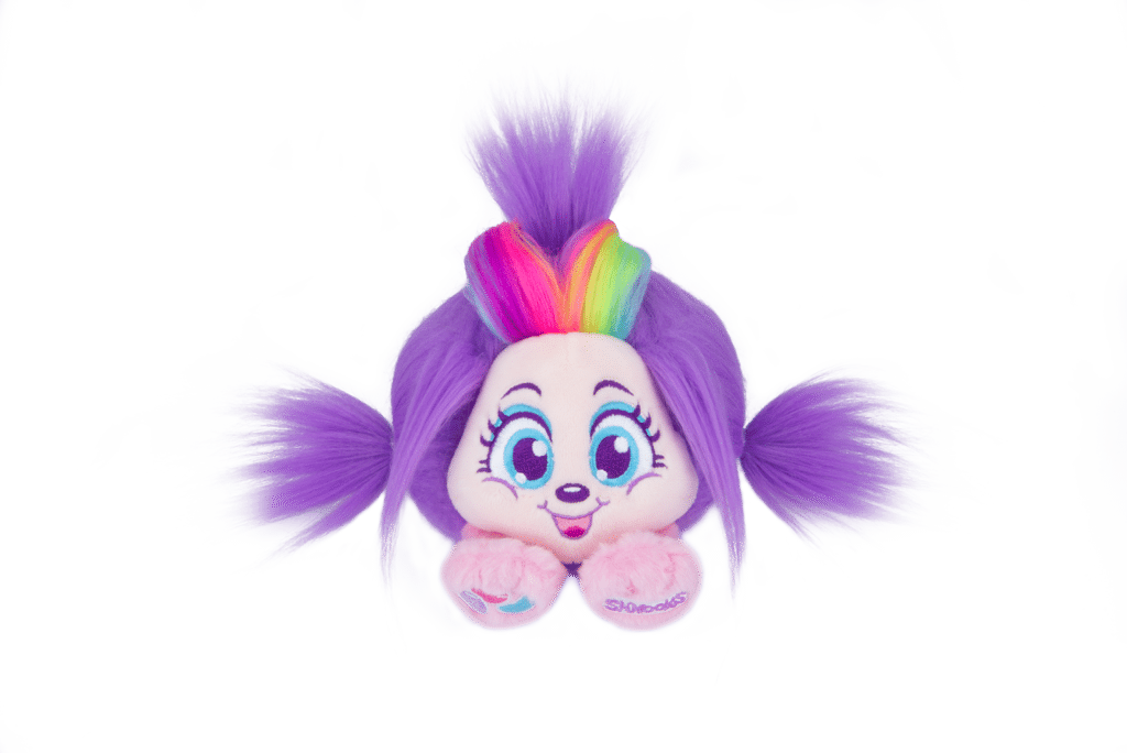 A soft toy with bright purple fur, large blue eyes, a smiling face, and two side ponytails. It has a tuft of rainbow-coloured hair on top and holds its small pink hands in front, set against a plain white background.