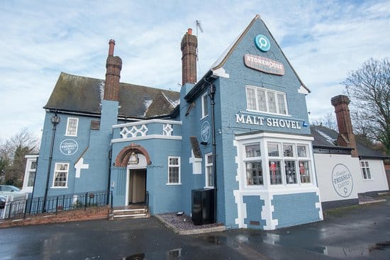 A large blue pub building with white trim, labelled "Malt Shovel" and "Stonehouse." It has tall brick chimneys, a gabled roof, and a white-framed bay window. The ground is wet, and leafless trees are visible in the background.
