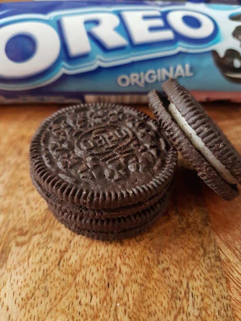 Three Oreo biscuits are stacked on a wooden surface, with one Oreo leaning against the stack. In the background, a partially visible blue packet labelled “Oreo Original” is slightly out of focus. The biscuits show the classic embossed design.