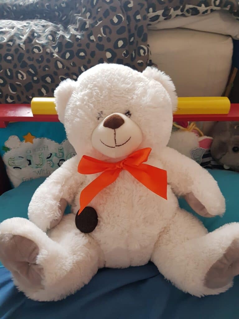 A fluffy white teddy bear with a red-orange ribbon bow round its neck sits on a blue surface. The bear has a brown nose, stitched smile, and grey paw pads. Behind it are cushions, one with stars and text, and a leopard-print blanket.