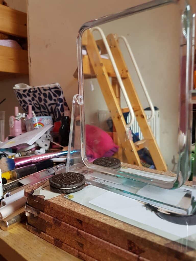 A cluttered wooden desk with a small glass mirror reflecting part of a bunk bed and a radiator. On the desk are Oreo biscuits, pens, make-up items, and a patterned pouch. The room appears busy and lived-in, with items scattered about.