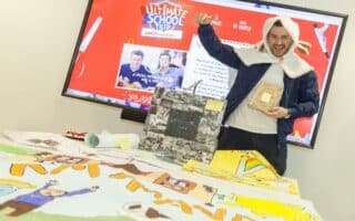 A smiling man wearing a hoodie with floppy ears poses excitedly in front of a screen displaying “The Ultimate School Trip.” He holds a trophy and stands behind a table full of colourful drawings and school projects, celebrating a competition or event.