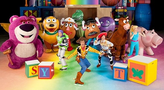 A group of performers dressed as Toy Story characters, including Woody, Buzz, Jessie, Bo Peep, and others, pose together on colourful, lit-up flooring. Large alphabet blocks and cartoon props decorate the cheerful, vibrant scene.
