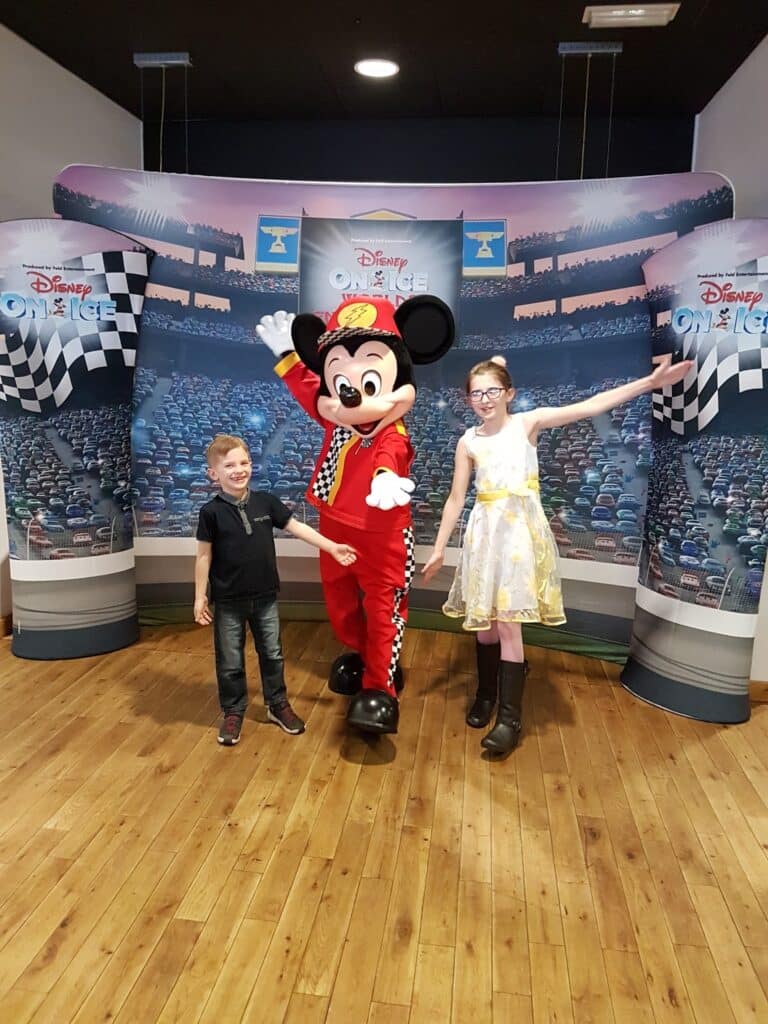 A smiling boy and girl pose with a person in a Mickey Mouse costume dressed as a racing car driver, standing on a wooden floor in front of a "Disney On Ice" backdrop with chequered flags and a stadium full of cars.