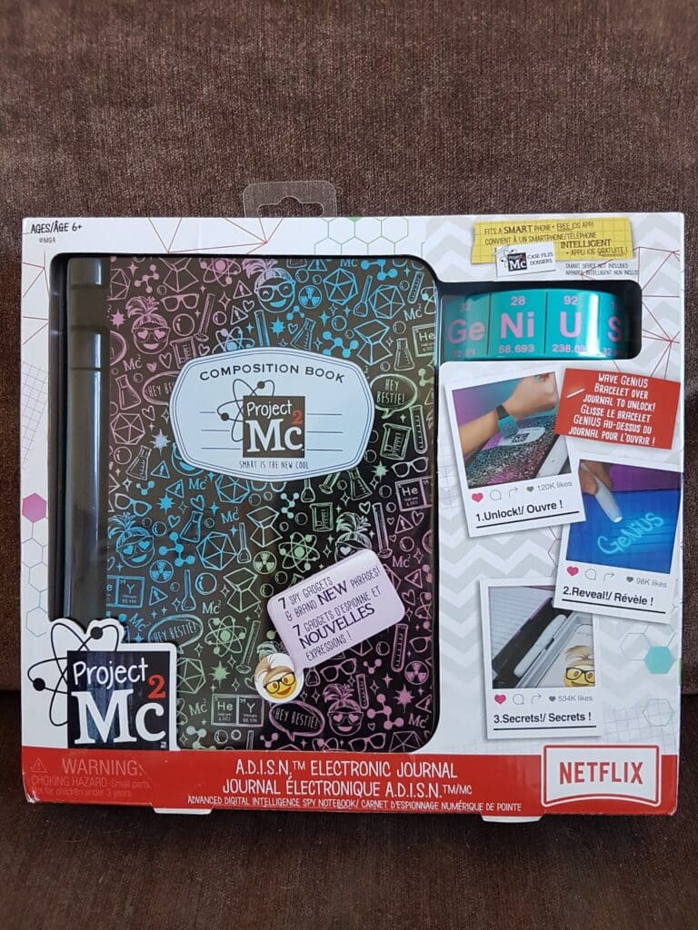 The image shows a boxed "Project Mc² A.D.I.S.N. Electronic Journal" toy. The packaging features a black, colourful journal, a purple pen, and accessories. The box is decorated with the Project Mc² logo and Netflix branding, and instructions for unlocking secrets.